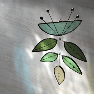 May include: A stained glass mobile featuring a teal flower and green leaves. The mobile is suspended from a wire and has a delicate, whimsical design.