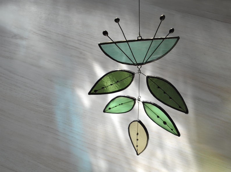 May include: A stained glass mobile featuring a teal flower and green leaves. The mobile is suspended from a wire and has a delicate, whimsical design.