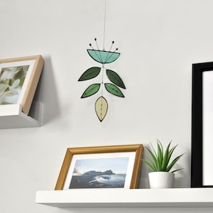 May include: A stained glass mobile with green and blue leaves hanging from a white wall. The mobile is hanging above a white shelf with a framed photo and a potted plant.