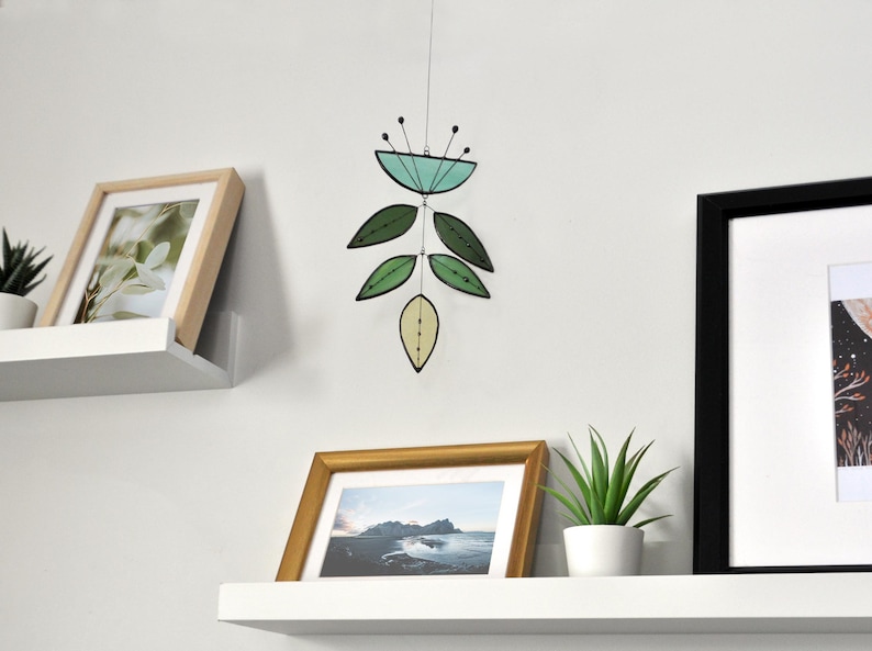 May include: A stained glass mobile with green and blue leaves hanging from a white wall. The mobile is hanging above a white shelf with a framed photo and a potted plant.