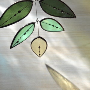 May include: A stained glass mobile with five leaves in varying shades of green and yellow. Each leaf has a dark outline and small, dark glass beads. The leaves are suspended by wire, creating a delicate, nature-inspired decoration for the home.