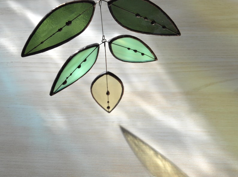 May include: A stained glass mobile with five leaves in varying shades of green and yellow. Each leaf has a dark outline and small, dark glass beads. The leaves are suspended by wire, creating a delicate, nature-inspired decoration for the home.