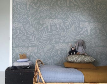 May include: A child's bedroom with a light blue wall featuring a white tiger and leaf pattern. A rattan bed with blue and gold bedding is in the foreground, with stuffed animals and pillows. A black trunk holds folded blankets.
