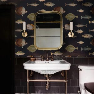Puede incluir: Interior de baño con lavabo blanco y grifería de latón, complementado con un espejo con marco de latón. Las paredes están decoradas con papel pintado azul oscuro con ilustraciones de peces. Azulejos negros y una puerta negra completan el diseño.