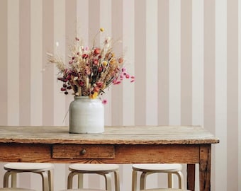 Op de afbeelding: Een rustieke houten tafel met een lade, met een witte keramische vaas gevuld met gedroogde bloemen. Zes kleine, lichtgekleurde houten krukken staan voor de tafel. De achtergrond is een gestreepte muur in roze en witte tinten.