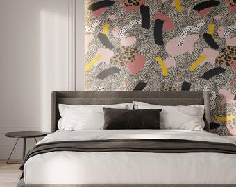 May include: A modern bedroom with a gray upholstered bed, white bedding, and a dark gray throw blanket. The headboard is against a wall with a colorful abstract wallpaper featuring pink, black, yellow, and leopard print patterns. A small side table is visible.