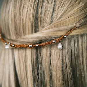 Könnte beinhalten: Ein braunes Makramee-Haaraccessoire mit silbernen Perlen und einem tropfenförmigen Anhänger aus Perlmutt. Das Haarband wird in blondem Haar getragen und verleiht einen Hauch von Bohème-Eleganz. Ideal für Hochzeiten oder besondere Anlässe.