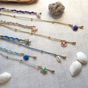 Puede incluir: Una colección de tobilleras con temática playera. Cada tobillera presenta un diseño diferente, incluyendo conchas marinas, estrellas de mar y perlas. Las tobilleras son de varios colores, como azul, morado y dorado. Las tobilleras se exhiben sobre una superficie blanca.