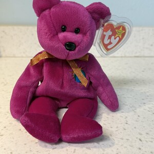 Puede incluir: Un oso de peluche magenta con una cinta dorada atada alrededor del cuello. El oso tiene una nariz negra, ojos negros pequeños y una etiqueta en forma de corazón. La etiqueta dice "Ty Beanie Baby".