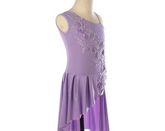 May include: A lavender dance dress with a one-shoulder design. The dress features floral embellishments on the bodice and a flowing, asymmetrical skirt. The dress is displayed on a mannequin against a white background.
