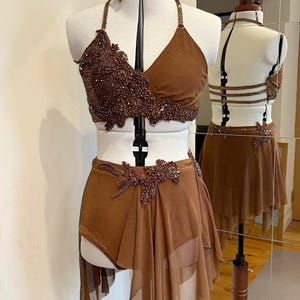 May include: A brown dance costume set on a mannequin, featuring a halter top, shorts with a skirt overlay, and a headpiece. The top and shorts are embellished with decorative beading. A second costume is visible in the mirror.