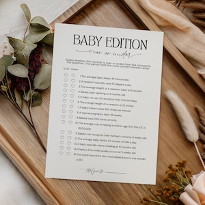 May include: A cream-colored baby shower game card titled "Baby Edition" with a list of questions. The card is on a wooden tray with decorative flowers and greenery. The game is "Over or Under".
