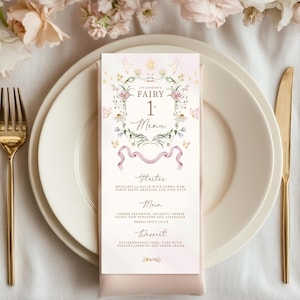 May include: A menu card with a floral design, featuring the text "ITS JASMINE'S FAIRY 1st Menu". The menu lists a starter, main course, and dessert. The card is placed on a white plate with gold cutlery.
