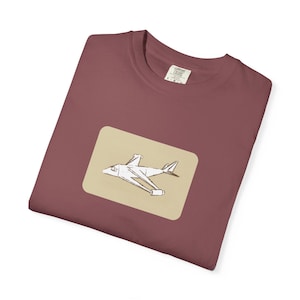 Könnte beinhalten: Ein weinrotes T-Shirt mit einem beigen Rechteck, das eine weiße Flugzeugillustration zeigt. Das Flugzeugdesign ist eine einfache Strichzeichnung. Das Shirt hat einen Rundhalsausschnitt und kurze Ärmel. Das Etikett auf dem Shirt lautet "COMFORT COLORS".
