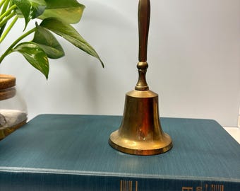 Może przedstawiać: Zabytkowy mosiężny dzwonek z długą rączką, spoczywający na turkusowej książce. Dzwonek ma klasyczny design z rozszerzoną podstawą i zaokrągloną górą. Uchwyt jest ciemniejszym odcieniem mosiądzu. Książka ma złote litery.