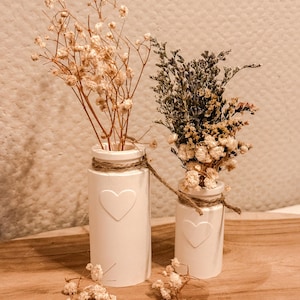 Könnte beinhalten: Zwei weiße Keramikvasen mit herzförmigen Ausschnitten, jede mit Trockenblumen. Die Vasen sind mit Bindfaden umwickelt und stehen auf einer Holzoberfläche. Eine Vase enthält beige Blüten, die andere eine Mischung aus Grün und Beige.