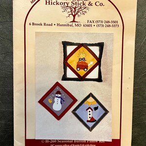May include: A craft kit for a seasonal button pillow. The kit includes a 14-inch square pillow with three interchangeable appliqued panels. The panels feature a snowman, a fall scene with a pumpkin wagon, and a birdhouse.