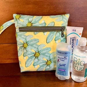 May include: A yellow floral zippered pouch with a handle, containing a blue deodorant stick labeled "Secret Aluminum Free Gentle on Skin", a blue bottle of eye drops labeled "Bausch + Lomb Biotrue", and a white tube of cream labeled "Isodine Pronamel".