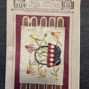 May include: A craft pattern titled "Little Stitches" featuring a floral design with a dark blue purse-like shape and colorful flowers. The pattern is on a cream-colored background with a burgundy border and decorative elements.