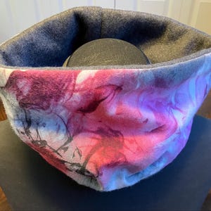 May include: A cozy neck warmer with a soft, gray fleece lining and a colorful, abstract print featuring shades of pink, purple, and blue.