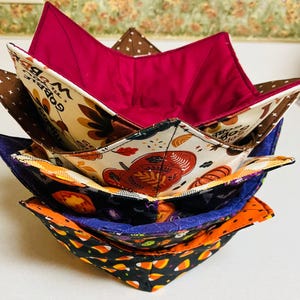 May include: Four fabric bowls in a stack. The top bowl is a solid burgundy color. The second bowl has a white background with brown, orange, and yellow pumpkins and leaves. The third bowl is a dark purple with a white and orange pattern. The bottom bowl is orange with a black and white candy corn pattern.