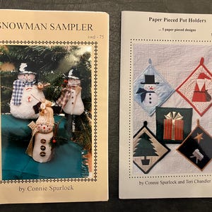 May include: Two craft pattern books. The first, "Snowman Sampler," features a framed image of three snowmen. The second, "Paper Pieced Pot Holders," displays five quilted pot holder designs with holiday themes. Both books are by different authors.