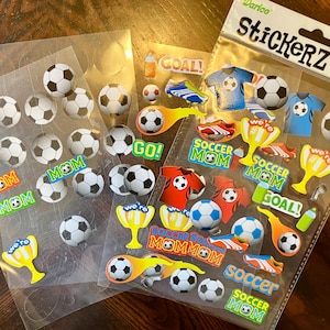 May include: A collection of soccer-themed stickers, featuring soccer balls, jerseys, trophies, and the words "GOAL" and "Soccer Mom." The stickers are in various colors and designs, perfect for decorating notebooks or crafts.