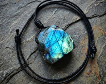 Puede incluir: Un collar con colgante de labradorita con un cordón negro. La piedra de labradorita es una mezcla de azul, verde y gris, con una estructura cristalina. El collar se muestra sobre una superficie de piedra gris.