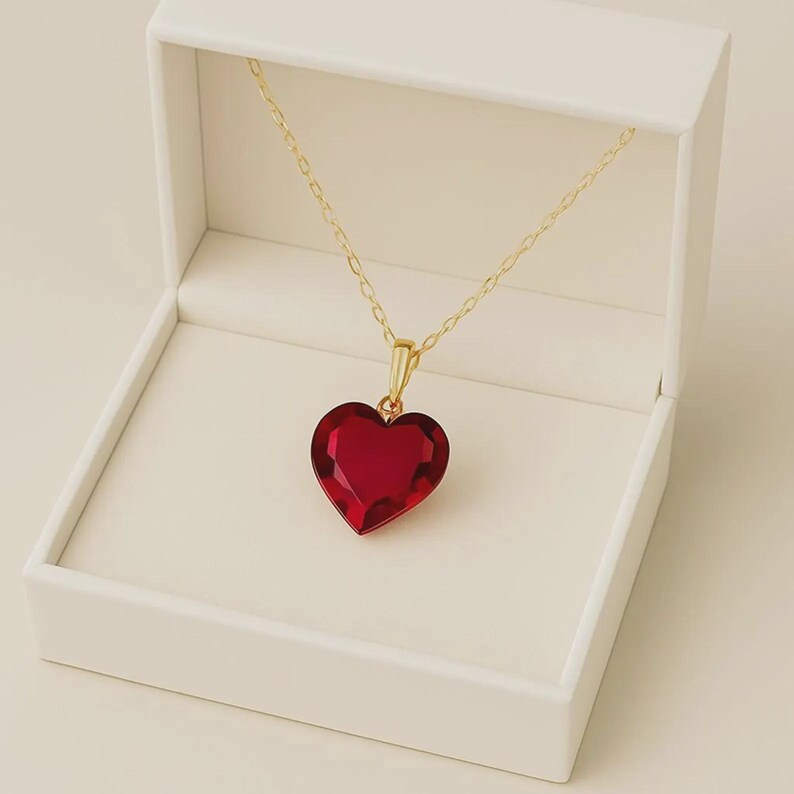 May include: A heart-shaped, ruby-red pendant necklace with a gold chain and clasp, presented in an open white jewelry box. The pendant is faceted, catching the light. The necklace is a romantic accessory.