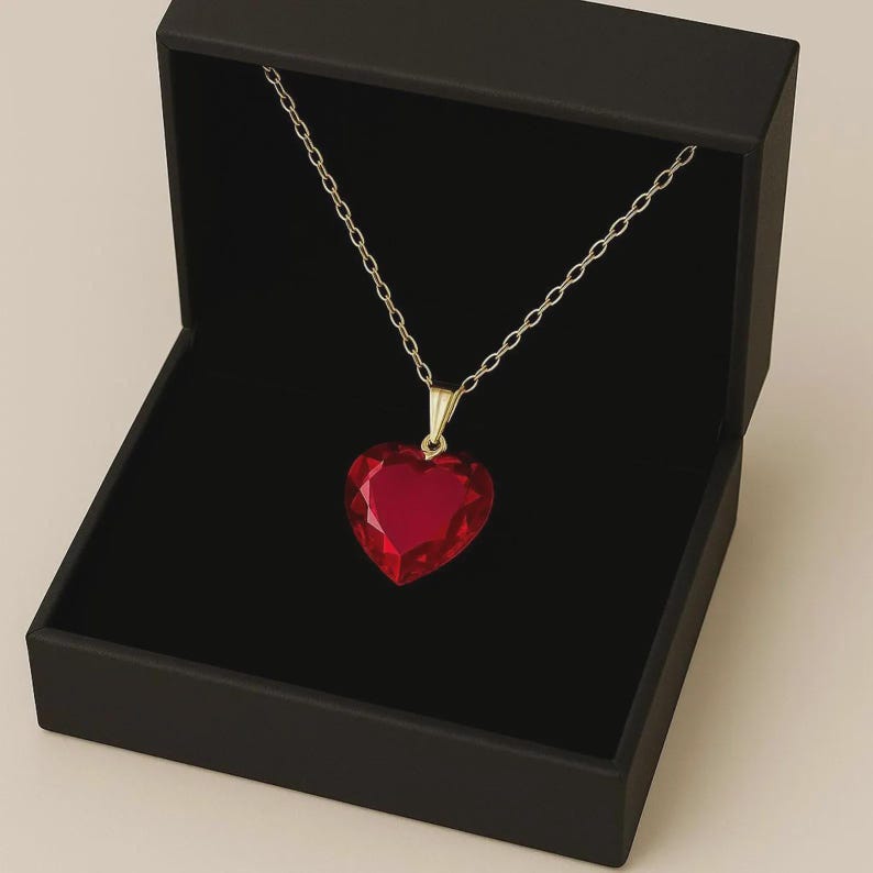 May include: A gold-toned necklace with a large, heart-shaped, faceted red pendant. The necklace is displayed in an open black jewelry box against a neutral background. The pendant is a deep, vibrant red.