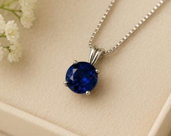 May include: A silver necklace with a round, deep blue gemstone pendant. The pendant is set in a four-prong setting and hangs from a delicate chain. The background is a soft, neutral color, with white flowers in the upper left corner.