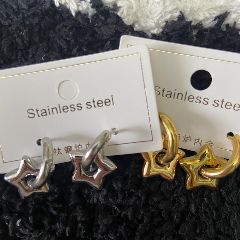 May include: Two pairs of stainless steel hoop earrings with star-shaped charms. One pair is silver-toned, and the other is gold-toned. Each pair is displayed on a white card with the words "Stainless steel".