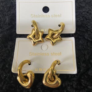 May include: Gold-colored stainless steel earrings. One pair features star-shaped pendants, and the other pair has heart-shaped pendants. The earrings are displayed on white cards with the text "Stainless steel".