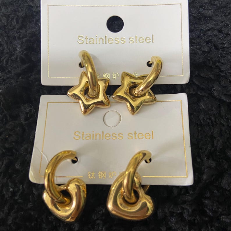 May include: Gold-colored stainless steel earrings. One pair features star-shaped pendants, and the other pair has heart-shaped pendants. The earrings are displayed on white cards with the text "Stainless steel".