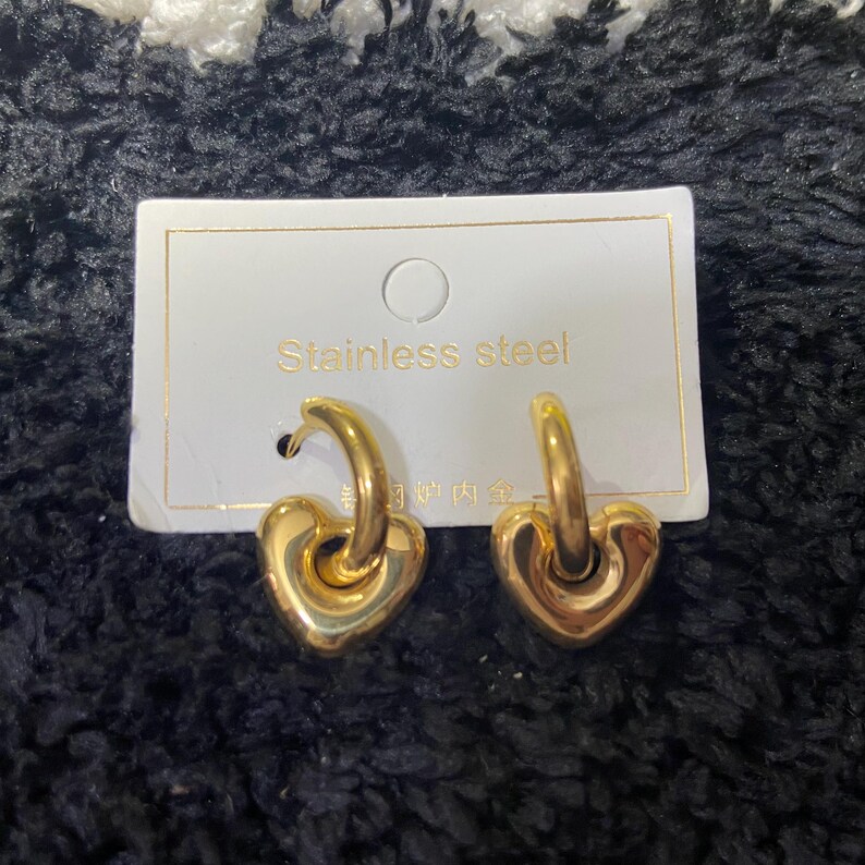 May include: A pair of gold-colored heart-shaped hoop earrings. The earrings are made of stainless steel and are displayed on a white card with the words "Stainless steel" printed on it.