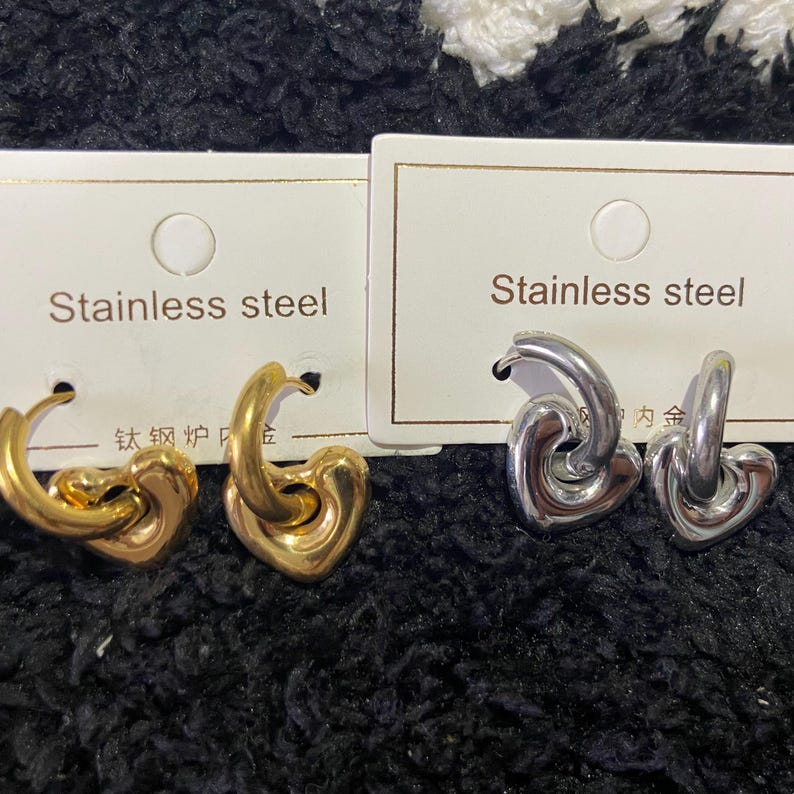 May include: Two pairs of heart-shaped earrings. One pair is gold-colored, the other is silver-colored. Each pair is displayed on a white card that reads "Stainless steel". The earrings are on a black, textured surface.