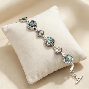 Puede incluir: Una pulsera de tono plateado con eslabones circulares y cuadrados alternos, cada uno incrustado con concha de abulón iridiscente. Los engastes de metal tienen un borde decorativo texturizado. La pulsera incluye un cierre de barra.
