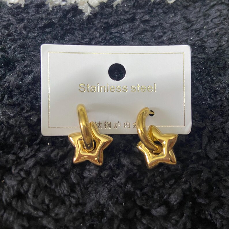 May include: A pair of gold-colored star-shaped earrings. The earrings are attached to a gold hoop. The earrings are on a white card with the words "Stainless Steel" printed on it.