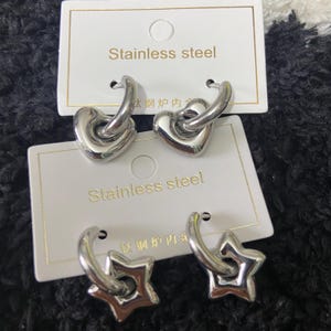 May include: Two pairs of stainless steel earrings. One pair features heart-shaped charms, and the other has star-shaped charms. The earrings are silver-colored and are displayed on white cards with the text "Stainless steel".