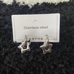 May include: A pair of silver-colored star-shaped hoop earrings. The earrings are made of stainless steel and are displayed on a white card with the words "Stainless steel" printed on it.