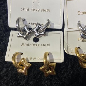 May include: Close-up of silver and gold-colored star-shaped hoop earrings. The earrings are displayed on white cards with the text "Stainless steel". The earrings have a shiny finish and a star design.