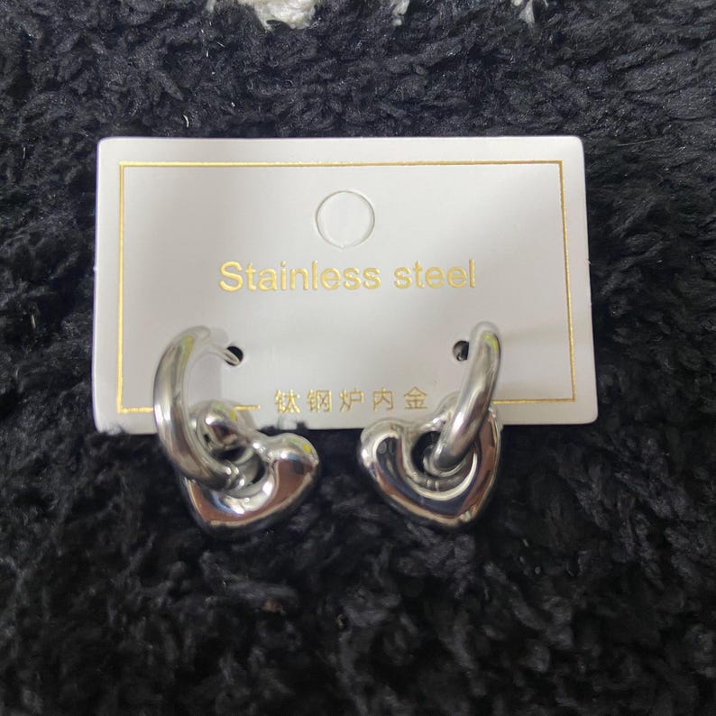 May include: A pair of stainless steel heart-shaped hoop earrings. The earrings are silver and have a polished finish. The earrings are displayed on a white card with the words "Stainless steel" printed on it.