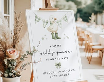 May include: A white sign on a wooden easel with a baby shower theme. The sign features a cartoon goose wearing green boots, a banner, and the text "A little silly goose is on the way!" and "Welcome to Ashley's Baby Shower."