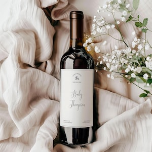 Puede incluir: Una botella de vino con una etiqueta personalizada que dice "Baby Thompson". La etiqueta también incluye el texto "Family Reserve" y "Due May 20XX". La botella oscura tiene una parte superior marrón y está sobre una tela de tono neutro y flores blancas.