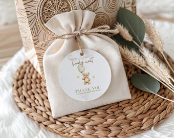 May include: A beige drawstring bag with a round tag that reads "We can bearly wait" with a teddy bear illustration. The bag is tied with twine and sits on a woven placemat, with decorative sprigs and a gift box in the background.