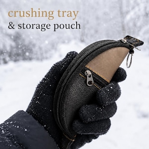 Puede incluir: Una bandeja de trituración y bolsa de almacenamiento en forma de media luna con exterior negro texturizado, panel de acento color canela y cierre de cremallera. El texto "crushing tray & storage pouch" aparece sobre un fondo nevado.