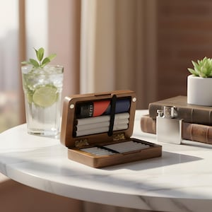 May include: A wooden cigarette case with a hinged lid, open to reveal cigarettes and a lighter. A glass of water with ice, lime, and mint sits nearby. A small succulent in a white pot rests on a stack of books.