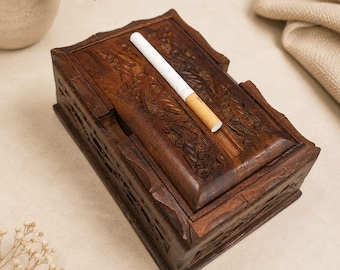May include: A rectangular dark brown wooden box featuring intricate carved leaf and vine patterns on its lid and sides. A single white cigarette with a tan filter rests diagonally across the top surface of the box, which has a hinged lid design.