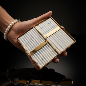 Puede incluir: Una pitillera rectangular de metal dorado abierta que muestra dos compartimentos interiores. Cada lado contiene una fila de cigarrillos blancos con marcas de filtro azules, sujetos por una fina barra metálica horizontal de color dorado.
