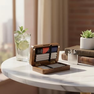 May include: A wooden cigarette case, open to reveal cigarettes and accessories, sits on a marble table. A glass of water with lime and a small succulent in a white pot are also on the table. A silver flask and books are in the background.